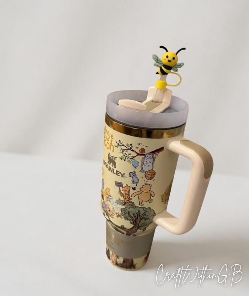 Puede incluir: Un termo de viaje Stanley beige con personajes de Winnie the Pooh, con borde y asa dorados. El termo tiene una tapa transparente y un adorno de pajita con forma de abeja amarilla. El termo est&aacute; decorado con ilustraciones de Winnie the Pooh y sus amigos.