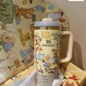 Puede incluir: Un termo de viaje Stanley con personajes de Winnie the Pooh en un esquema de color amarillo y crema. El termo tiene un asa y tapa blancas. El texto "Winnie the Pooh" y "Stanley" son visibles.