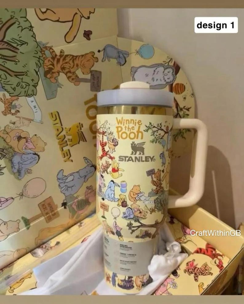 Puede incluir: Un termo de viaje Stanley con personajes de Winnie the Pooh en un esquema de color amarillo y crema. El termo tiene un asa y tapa blancas. El texto "Winnie the Pooh" y "Stanley" son visibles.