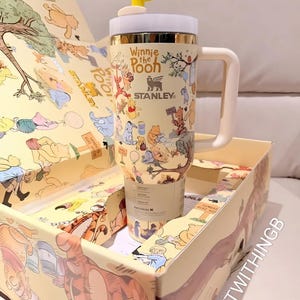 Puede incluir: Un vaso Stanley con el tema de Winnie the Pooh, con un asa amarilla y blanca y una tapa con forma de abeja. El vaso est&aacute; en una caja a juego. El vaso tiene las palabras "Winnie the Pooh" y el logotipo de Stanley.