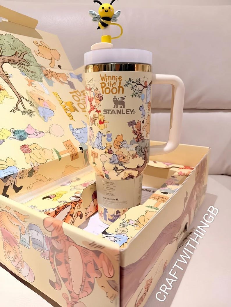 Puede incluir: Un vaso Stanley con el tema de Winnie the Pooh, con un asa amarilla y blanca y una tapa con forma de abeja. El vaso est&aacute; en una caja a juego. El vaso tiene las palabras "Winnie the Pooh" y el logotipo de Stanley.