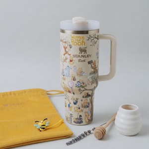 Puede incluir: Un termo de viaje Stanley beige con ilustraciones de Winnie the Pooh. El termo tiene una tapa y un asa blancas, con el texto "Winnie the Pooh" y "Stanley Beth". Tambi&eacute;n hay una bolsa amarilla, un dije de abeja, un cuchar&oacute;n de miel y un tarro blanco peque&ntilde;o.