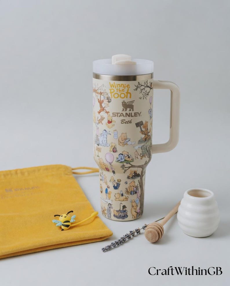 Puede incluir: Un termo de viaje Stanley beige con ilustraciones de Winnie the Pooh. El termo tiene una tapa y un asa blancas, con el texto "Winnie the Pooh" y "Stanley Beth". Tambi&eacute;n hay una bolsa amarilla, un dije de abeja, un cuchar&oacute;n de miel y un tarro blanco peque&ntilde;o.