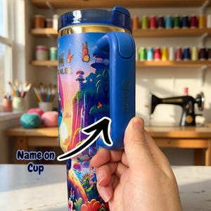 Puede incluir: Un termo de viaje azul y dorado con un colorido dise&ntilde;o de paisaje de fantas&iacute;a con mariposas y un castillo. El termo tiene un asa azul con el nombre "Storm" impreso. El texto "Dark Blue Design" es visible.