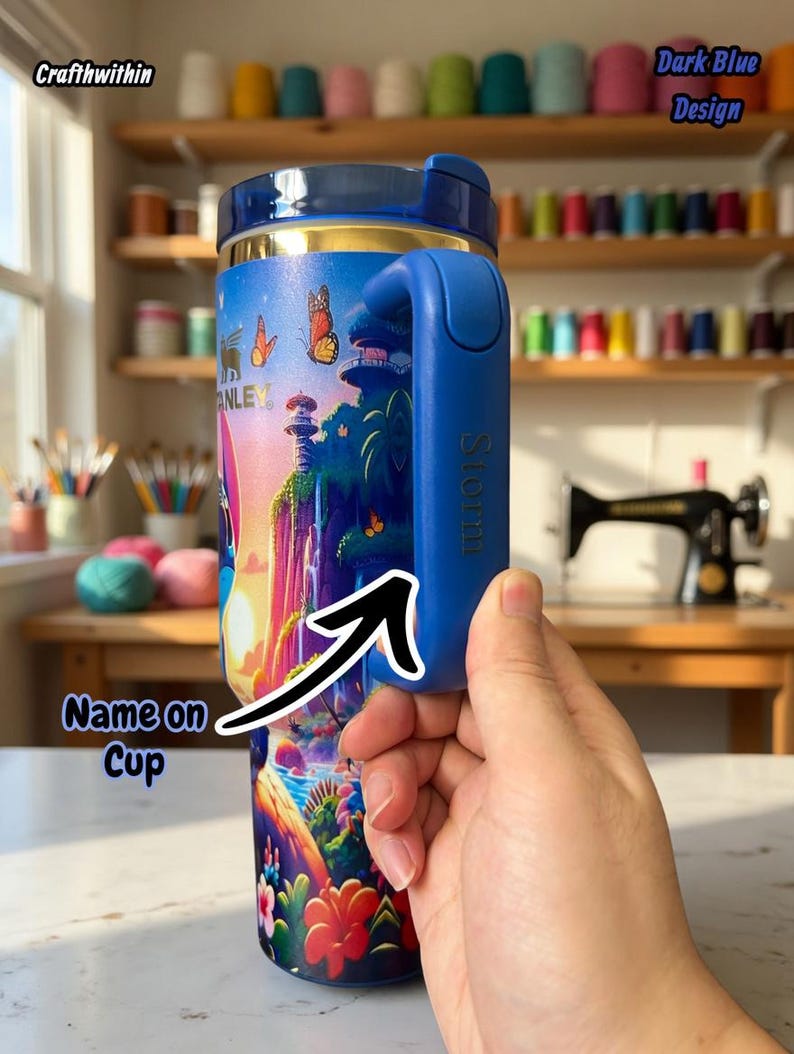 Puede incluir: Un termo de viaje azul y dorado con un colorido dise&ntilde;o de paisaje de fantas&iacute;a con mariposas y un castillo. El termo tiene un asa azul con el nombre "Storm" impreso. El texto "Dark Blue Design" es visible.