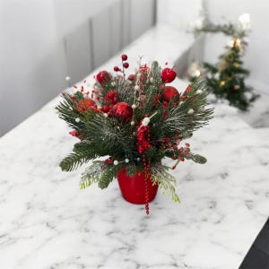 以下が含まれることがあります： 赤いポットに入ったお祝いのクリスマスアレンジメント。緑の松の枝、赤いオーナメント、ベリー、白いアクセントが特徴です。ポットは白い大理石の表面に置かれ、背景には小さなクリスマスツリーがあります。