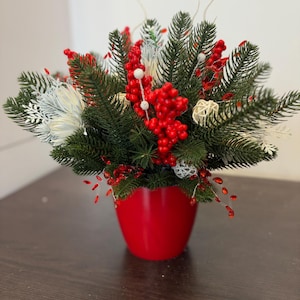 May include: A festive arrangement of evergreen branches, red berries, and white accents in a glossy red pot. The holiday-themed centerpiece features a mix of textures and colors, perfect for seasonal decor.
