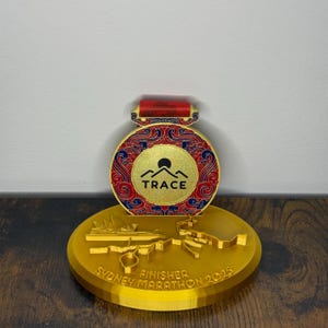 Peut inclure: Médaille de finisher du marathon de Sydney 2023, dorée. La médaille présente un motif rond avec des motifs rouges et bleus, le mot "TRACE" et un logo de montagne. La médaille est sur une base dorée.