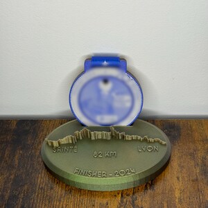 Peut inclure: Une médaille bleue et blanche avec un dessus bleu, posée sur une base ovale verte. La base porte les inscriptions "SAINTE", "82 KM" et "LYON", avec "FINISHER - 2024" en dessous. La médaille est probablement pour une course.