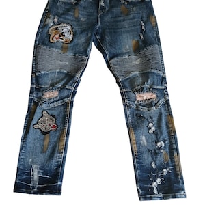 May include: Blue denim jeans with distressed details, patches, and decorative embellishments. Features a tiger patch on the left leg and a bear patch on the right leg. The jeans have a faded wash and ripped details.