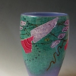May include: A glass vase with a tapered cylindrical shape, featuring a gradient from a light purple base to a textured teal upper section. The surface is decorated with abstract shapes in lavender, pink, and dark brown lines across the teal area.