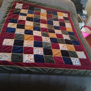 May include: A patchwork quilt with a grid pattern of various colored squares. The squares include burgundy, navy, gold, green, and patterned fabrics. The quilt has a burgundy border and a green velvet edge.