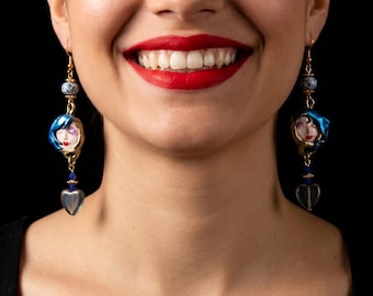 Könnte beinhalten: Hängeohrringe mit einer zentralen, gesichtsförmigen Perle mit blauem Haar, akzentuiert durch eine kleine graue Sprenkelperle darüber und eine durchscheinende herzförmige Perle darunter, verbunden durch goldfarbene Metallelemente und blaue Kristalle.