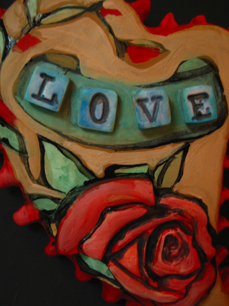 K&ouml;nnte beinhalten: Eine herzf&ouml;rmige Keramikskulptur mit einer roten Rose und dem Wort "LOVE" in blauen Fliesen.