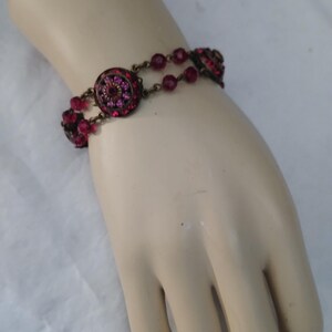 Puede incluir: Una pulsera de estilo vintage con un dise&ntilde;o circular central y peque&ntilde;as cuentas redondas de color rosa intenso. La pulsera tiene un acabado de lat&oacute;n antiguo, con el dise&ntilde;o central incrustado con cristales rosas m&aacute;s peque&ntilde;os.
