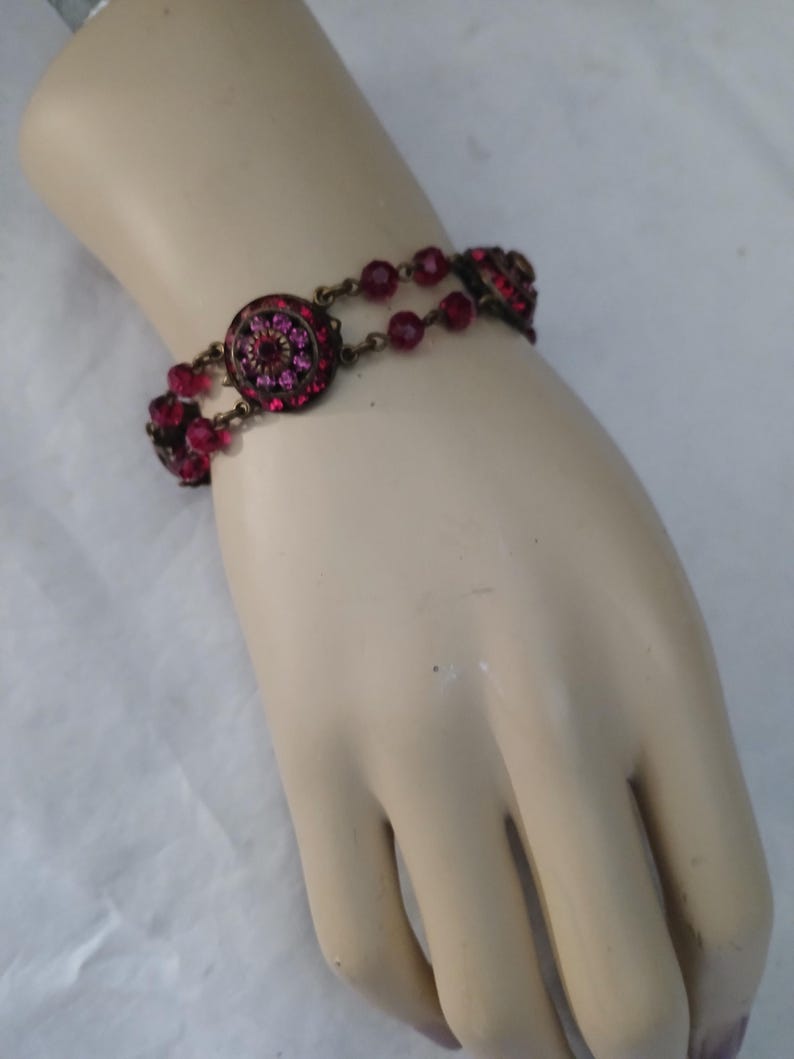 Puede incluir: Una pulsera de estilo vintage con un dise&ntilde;o circular central y peque&ntilde;as cuentas redondas de color rosa intenso. La pulsera tiene un acabado de lat&oacute;n antiguo, con el dise&ntilde;o central incrustado con cristales rosas m&aacute;s peque&ntilde;os.