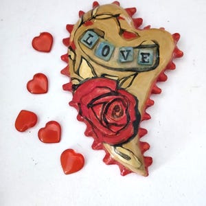K&ouml;nnte beinhalten: Ein herzf&ouml;rmiger Keramik-Anh&auml;nger mit einer roten Rose und dem Wort "LOVE" in blauen und wei&szlig;en Fliesen. Das Herz ist mit einem braunen und roten Rankenmuster verziert und hat einen gezackten Rand.