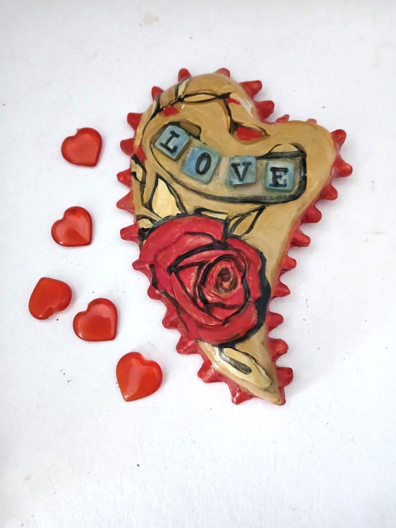 K&ouml;nnte beinhalten: Ein herzf&ouml;rmiger Keramik-Anh&auml;nger mit einer roten Rose und dem Wort "LOVE" in blauen und wei&szlig;en Fliesen. Das Herz ist mit einem braunen und roten Rankenmuster verziert und hat einen gezackten Rand.