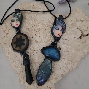 May include: Two handmade necklaces with black cords. One features a face pendant with a gear and a black crystal. The other has a face pendant and a blue and green stone. The pendants are set in black metal.