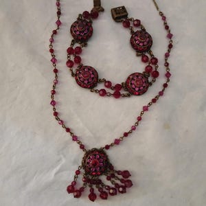 Puede incluir: Un conjunto de joyas vintage con collar y pulsera. El collar tiene un colgante redondo con cristales rosas y rojos y cuentas colgantes. La pulsera tiene tres medallones redondos con incrustaciones de cristal. Ambas piezas tienen un engaste de metal color bronce.