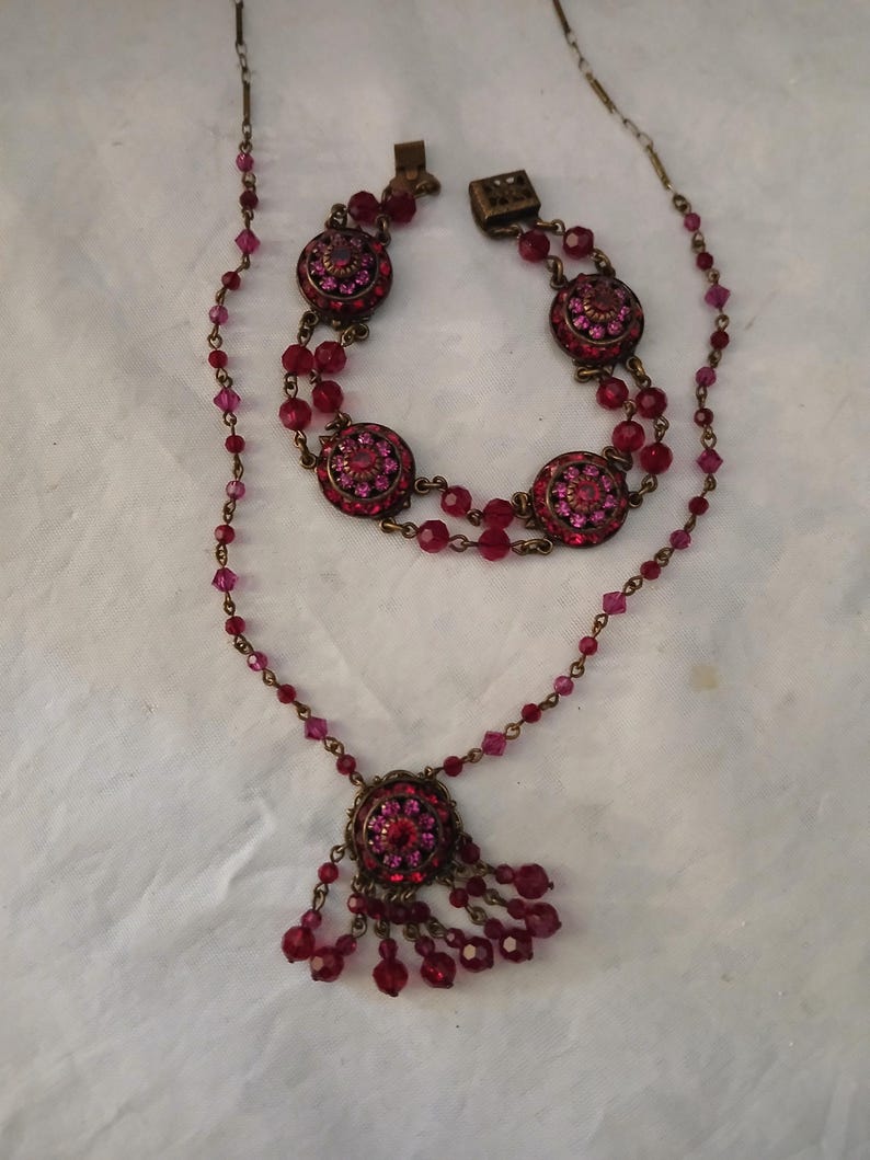 Puede incluir: Un conjunto de joyas vintage con collar y pulsera. El collar tiene un colgante redondo con cristales rosas y rojos y cuentas colgantes. La pulsera tiene tres medallones redondos con incrustaciones de cristal. Ambas piezas tienen un engaste de metal color bronce.