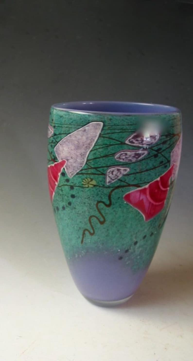 May include: A tapered glass vase featuring a vibrant teal and green speckled body with a solid lavender base and interior. The exterior is decorated with abstract purple shapes, bright pink conical forms, and thin dark brown wavy lines.
