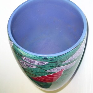 May include: A cylindrical glass vase with a smooth light blue interior rim and a deep periwinkle blue interior. The exterior features a mottled green surface decorated with abstract white, purple, and red geometric shapes and thin black lines.