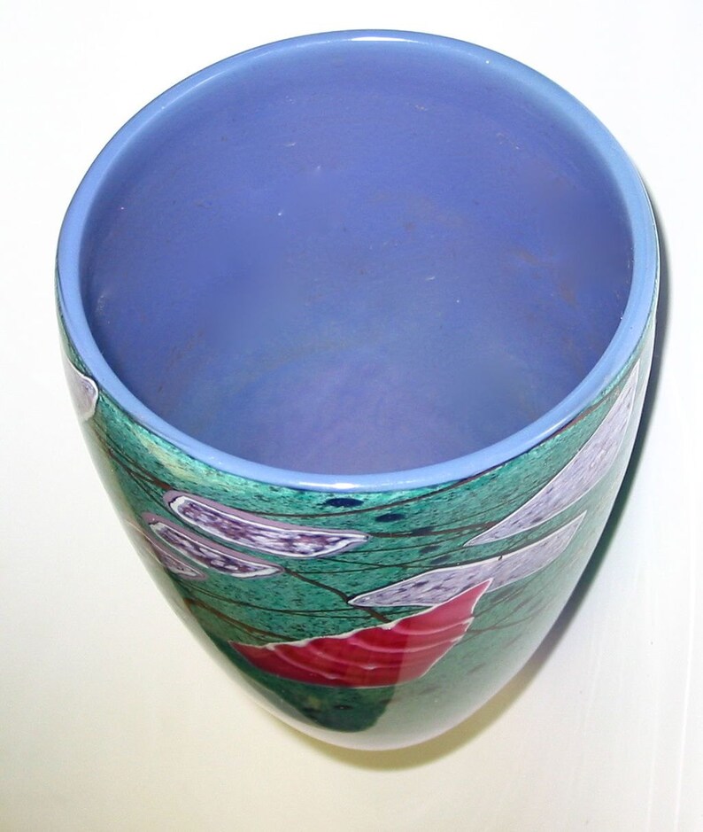 May include: A cylindrical glass vase with a smooth light blue interior rim and a deep periwinkle blue interior. The exterior features a mottled green surface decorated with abstract white, purple, and red geometric shapes and thin black lines.