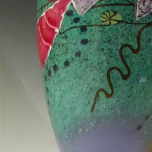 May include: A glass vase featuring a speckled green upper section and a solid light purple base. The surface is decorated with abstract shapes in bright pink and light purple, accented by thin dark brown wavy lines and small dark dots.