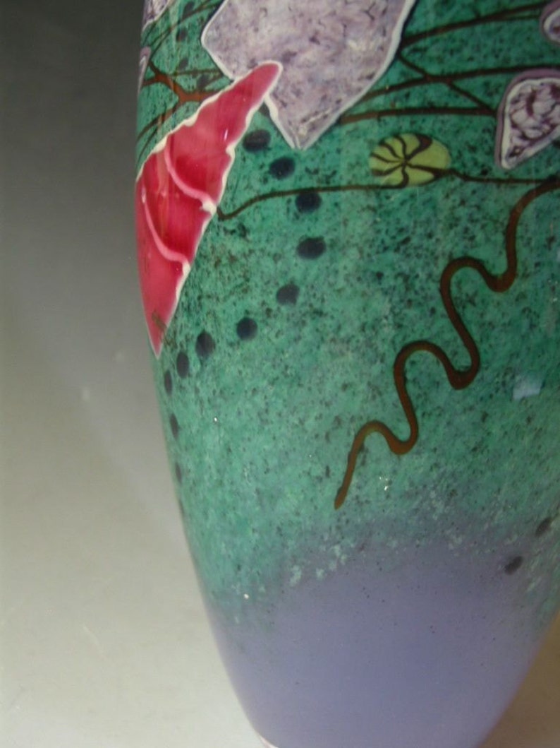 May include: A glass vase featuring a speckled green upper section and a solid light purple base. The surface is decorated with abstract shapes in bright pink and light purple, accented by thin dark brown wavy lines and small dark dots.