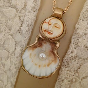 May include: A gold necklace with a pendant featuring two faces sculpted in white and brown, set inside a white seashell with a pearl in the center.