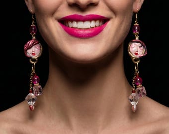 Könnte beinhalten: Ein Paar lange Ohrhänger mit einem skulpturalen Gesichtsanhänger in Pink-Rot, akzentuiert durch einen goldfarbenen Metallhaken, eine durchscheinende rosa Perle und eine klare facettierte Kristallperle an einer feinen goldfarbenen Kette.