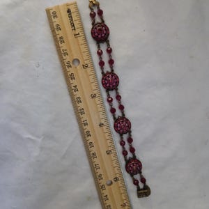 Puede incluir: Un brazalete vintage con tres elementos circulares con incrustaciones de joyas de color magenta, conectados por cuentas magenta m&aacute;s peque&ntilde;as. El brazalete mide aproximadamente 18 cm de largo. Una regla de madera est&aacute; al lado del brazalete.