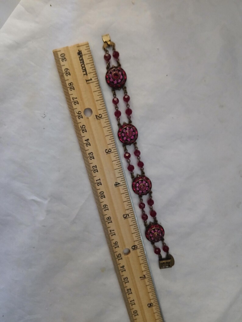 Puede incluir: Un brazalete vintage con tres elementos circulares con incrustaciones de joyas de color magenta, conectados por cuentas magenta m&aacute;s peque&ntilde;as. El brazalete mide aproximadamente 18 cm de largo. Una regla de madera est&aacute; al lado del brazalete.
