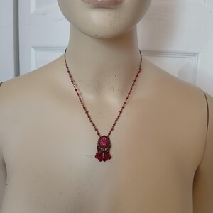 Puede incluir: Un collar de estilo vintage con una cadena de metal oscuro y un colgante con una piedra preciosa rosa central rodeada de piedras rosas m&aacute;s peque&ntilde;as. El colgante tiene cuentas rosas colgantes. El collar se muestra en un maniqu&iacute;.