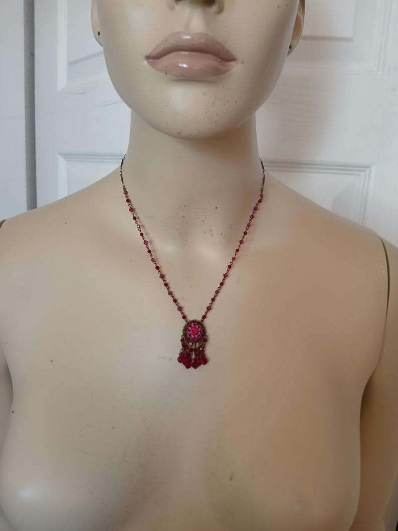 Puede incluir: Un collar de estilo vintage con una cadena de metal oscuro y un colgante con una piedra preciosa rosa central rodeada de piedras rosas m&aacute;s peque&ntilde;as. El colgante tiene cuentas rosas colgantes. El collar se muestra en un maniqu&iacute;.