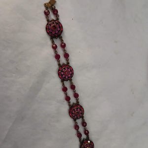Puede incluir: Un brazalete vintage con engastes redondos y ornamentados adornados con brillantes piedras preciosas rosas. El brazalete est&aacute; conectado por hilos de cuentas rosas m&aacute;s peque&ntilde;as a juego. El metal tiene un c&aacute;lido tono dorado antiguo y el cierre es visible en los extremos.