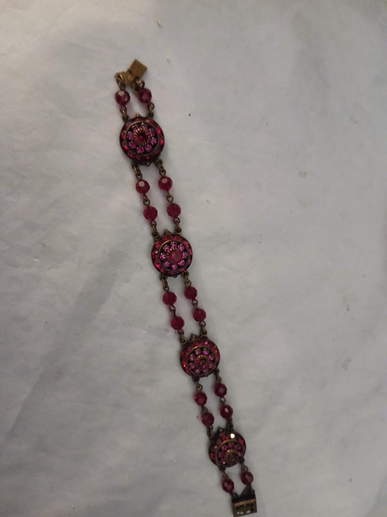 Puede incluir: Un brazalete vintage con engastes redondos y ornamentados adornados con brillantes piedras preciosas rosas. El brazalete est&aacute; conectado por hilos de cuentas rosas m&aacute;s peque&ntilde;as a juego. El metal tiene un c&aacute;lido tono dorado antiguo y el cierre es visible en los extremos.