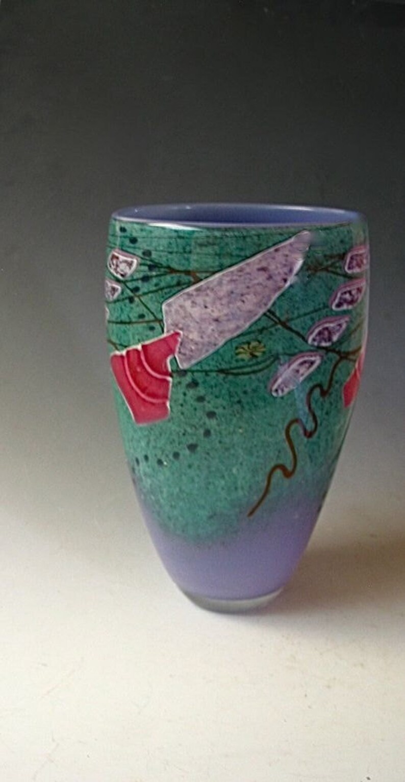 May include: A glass vase with a tapered cylindrical shape, featuring a gradient from a light purple base to a textured teal upper section. The surface is decorated with abstract shapes in lavender, pink, and dark brown lines across the teal area.