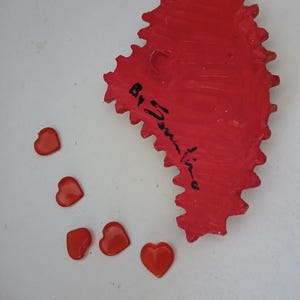 K&ouml;nnte beinhalten: Ein rotes, herzf&ouml;rmiges Keramikornament mit einem gezackten Rand und einer schwarzen Signatur, die "By Janine" lautet. F&uuml;nf kleine, rote, herzf&ouml;rmige Dekorationen sind um das Ornament herum verstreut.