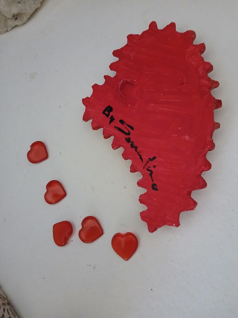 K&ouml;nnte beinhalten: Ein rotes, herzf&ouml;rmiges Keramikornament mit einem gezackten Rand und einer schwarzen Signatur, die "By Janine" lautet. F&uuml;nf kleine, rote, herzf&ouml;rmige Dekorationen sind um das Ornament herum verstreut.