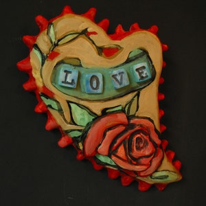 K&ouml;nnte beinhalten: Ein Keramik-Herz-Anh&auml;nger mit roter und brauner Glasur. Das Herz hat eine stilisierte Rose und gr&uuml;ne Bl&auml;tter. Das Wort "LOVE" ist in blauen und wei&szlig;en Fliesen geschrieben.