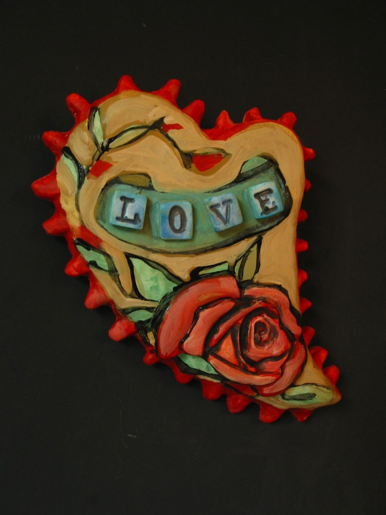 K&ouml;nnte beinhalten: Ein Keramik-Herz-Anh&auml;nger mit roter und brauner Glasur. Das Herz hat eine stilisierte Rose und gr&uuml;ne Bl&auml;tter. Das Wort "LOVE" ist in blauen und wei&szlig;en Fliesen geschrieben.