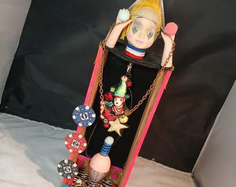 Könnte beinhalten: Eine skurrile Mixed-Media-Skulptur mit einem Puppenkopf mit bemaltem Clown-Make-up, einem Clown-Anhänger und einem federbelasteten Ei. Die Skulptur besteht aus Holz, Metall und Stoff und ist mit bunten Perlen, Pailletten und Pokerchips verziert. Die Basis der Skulptur ist mit einem Leopardenmusterstoff verziert und das Wort "JUMP" ist in Holzbuchstaben geschrieben.