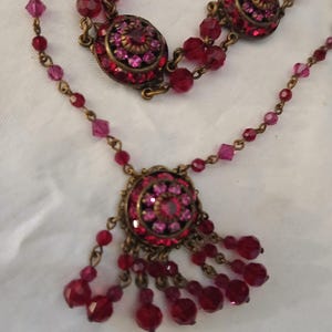Puede incluir: Un collar de estilo vintage con un dise&ntilde;o en capas. El collar presenta m&uacute;ltiples hilos de cuentas rojas y rosas, con colgantes circulares adornados con cristales rojos y rosas. El metal tiene un acabado bronce.
