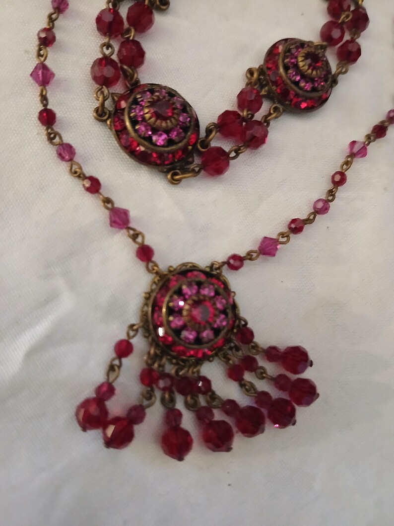 Puede incluir: Un collar de estilo vintage con un dise&ntilde;o en capas. El collar presenta m&uacute;ltiples hilos de cuentas rojas y rosas, con colgantes circulares adornados con cristales rojos y rosas. El metal tiene un acabado bronce.