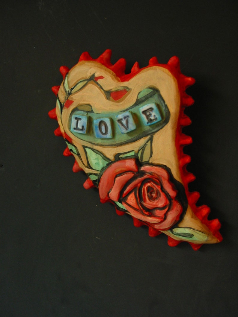 K&ouml;nnte beinhalten: Ein herzf&ouml;rmiger Keramik-Anh&auml;nger mit einer roten und wei&szlig;en Rose und dem Wort "LOVE" in bunten Fliesen.