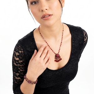 Puede incluir: Una mujer con un vestido de encaje negro y un conjunto de joyas a juego. El conjunto incluye un collar con colgante y una pulsera, ambos con piedras preciosas rojas. Fondo blanco.