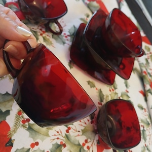 May include: Set of ruby red glass teacups and saucers. The cups have a rounded square shape with a handle. The saucers are square. The cups and saucers are arranged on a festive tablecloth with a holly and cardinal pattern.