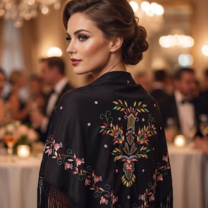 May include: A black shawl featuring intricate multi-colored floral embroidery on the back and along the edges. The garment is finished with long black fringe detailing along the bottom hem, creating a textured appearance across the fabric surface.